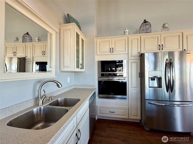 kitchen with dark wood-type flooring, cream cabinetry, a sink, stainless steel appliances, and glass insert cabinets