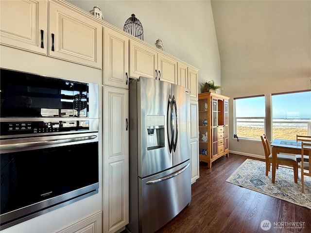 kitchen with dark wood-style floors, baseboards, cream cabinetry, appliances with stainless steel finishes, and a towering ceiling