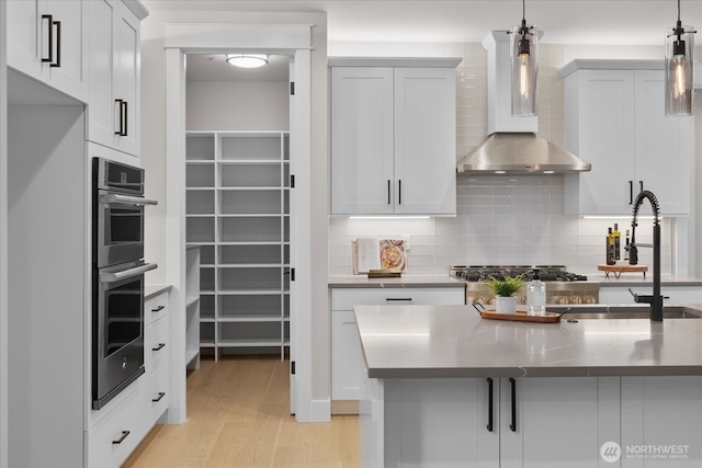 kitchen with light wood-style floors, decorative light fixtures, wall chimney range hood, double oven, and backsplash