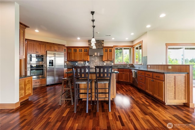 kitchen featuring range hood, a breakfast bar area, decorative backsplash, appliances with stainless steel finishes, and brown cabinetry