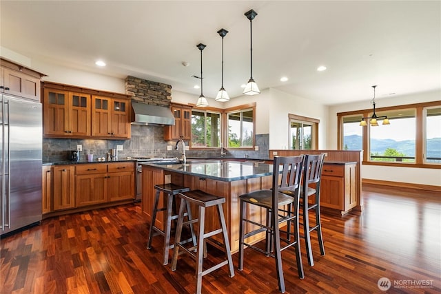 kitchen featuring premium appliances, brown cabinets, tasteful backsplash, a kitchen island with sink, and wall chimney exhaust hood