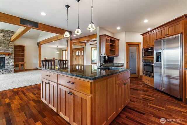 kitchen with dark stone counters, open floor plan, stainless steel appliances, a stone fireplace, and a sink