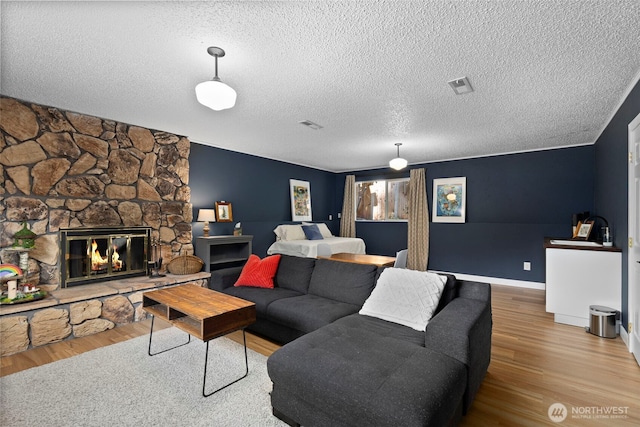 living room featuring wood finished floors, visible vents, baseboards, a stone fireplace, and a textured ceiling