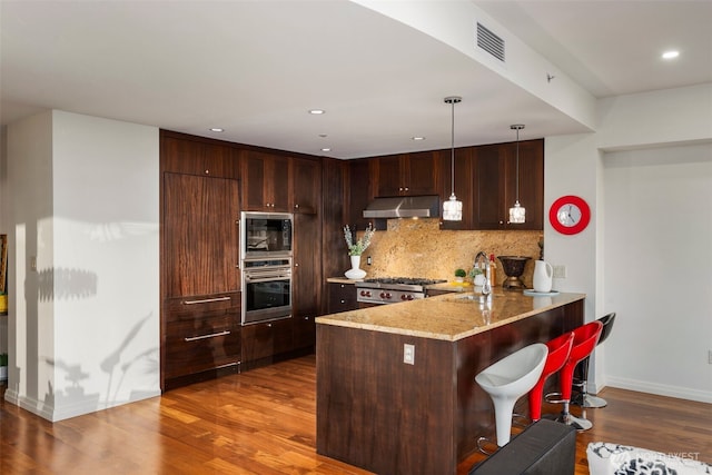 kitchen featuring backsplash, appliances with stainless steel finishes, a peninsula, wood finished floors, and under cabinet range hood