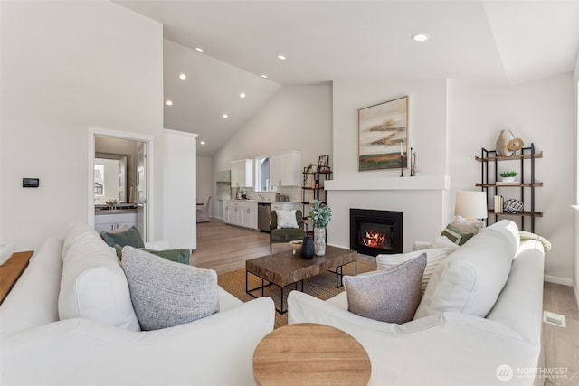 living area featuring visible vents, a glass covered fireplace, light wood-type flooring, high vaulted ceiling, and recessed lighting