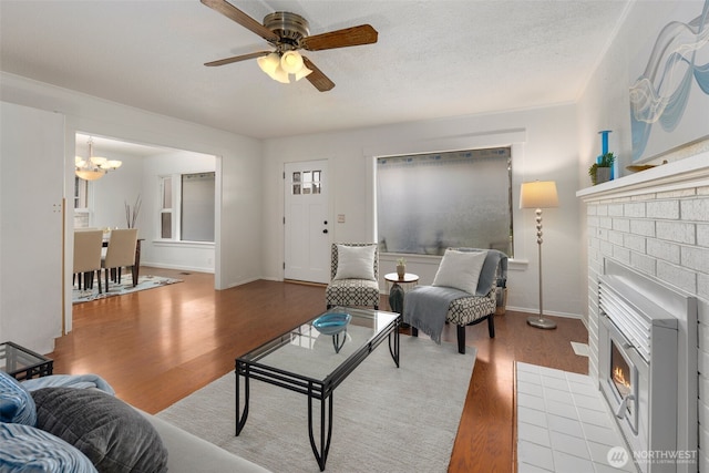 living room featuring a textured ceiling, ceiling fan with notable chandelier, wood finished floors, baseboards, and a brick fireplace