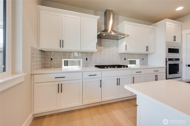 kitchen featuring stainless steel appliances, white cabinets, wall chimney range hood, light wood-type flooring, and backsplash