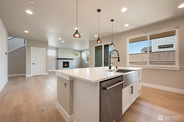 kitchen featuring a warm lit fireplace, a kitchen island with sink, a sink, dishwasher, and light wood finished floors