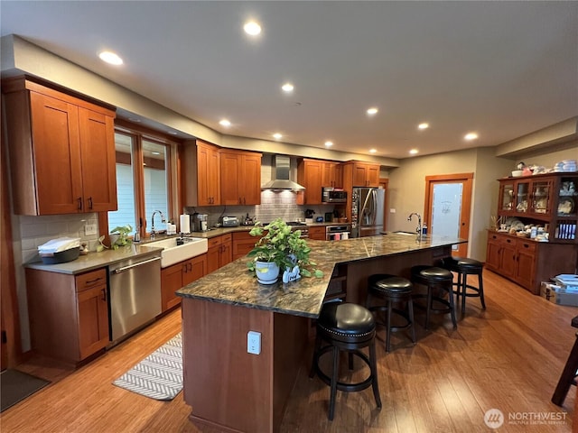 kitchen with stainless steel appliances, a sink, light wood-type flooring, wall chimney exhaust hood, and a kitchen bar