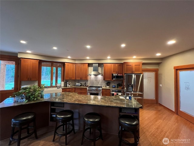 kitchen featuring a kitchen bar, backsplash, light wood-style flooring, appliances with stainless steel finishes, and wall chimney exhaust hood