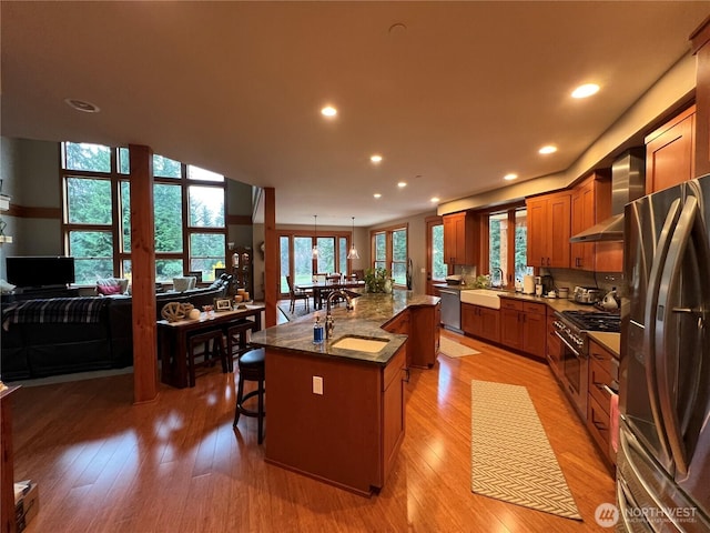 kitchen with stainless steel appliances, a sink, light wood-style floors, wall chimney range hood, and brown cabinetry