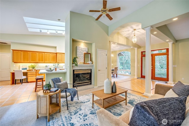 living area featuring high vaulted ceiling, light tile patterned flooring, a ceiling fan, a glass covered fireplace, and decorative columns