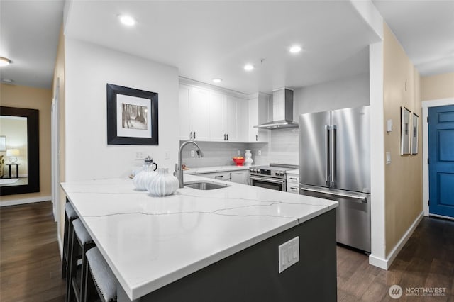 kitchen featuring appliances with stainless steel finishes, light stone counters, a peninsula, wall chimney range hood, and a sink