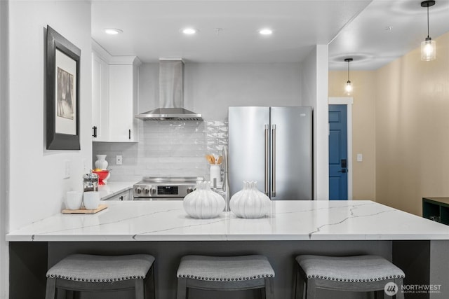 kitchen with tasteful backsplash, white cabinets, appliances with stainless steel finishes, light stone countertops, and wall chimney range hood