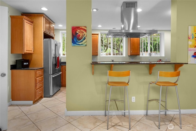 kitchen featuring light tile patterned floors, island range hood, stainless steel fridge with ice dispenser, dark countertops, and a kitchen bar