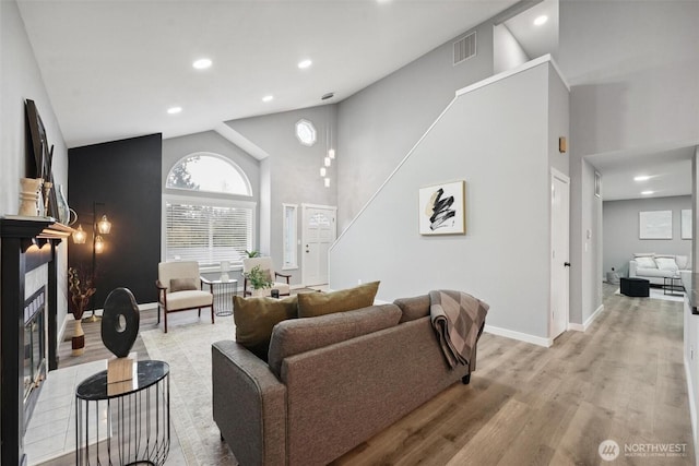 living area with visible vents, light wood-style flooring, a glass covered fireplace, high vaulted ceiling, and baseboards