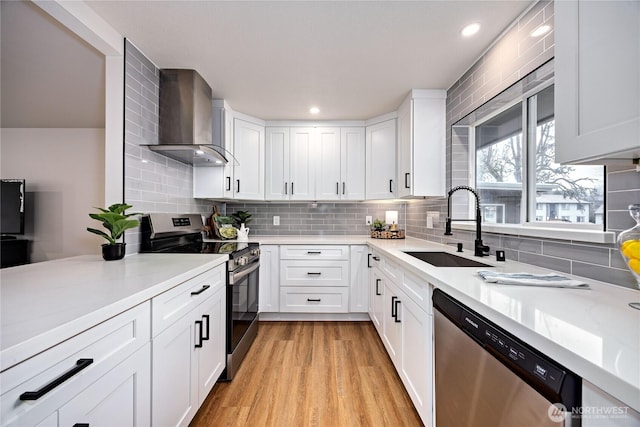 kitchen featuring appliances with stainless steel finishes, light wood-style floors, white cabinetry, a sink, and wall chimney exhaust hood