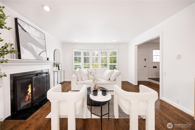 living area featuring a warm lit fireplace, baseboards, dark wood-style flooring, crown molding, and recessed lighting