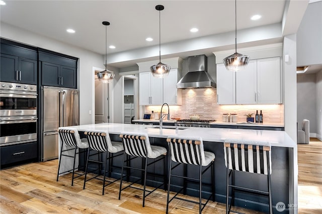 kitchen with light wood finished floors, light countertops, stainless steel appliances, wall chimney range hood, and a sink