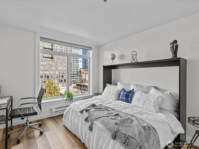 bedroom with a baseboard heating unit, a view of city, wood finished floors, and baseboards