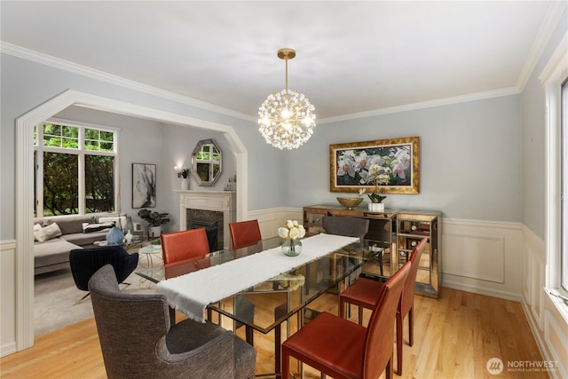 dining area with a wainscoted wall, light wood-style flooring, a premium fireplace, ornamental molding, and an inviting chandelier