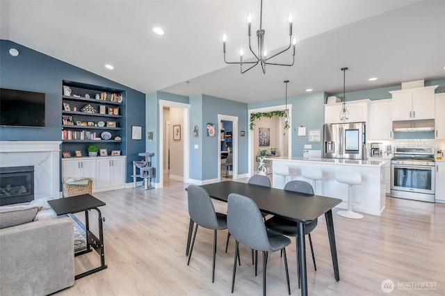 dining area featuring a premium fireplace, built in features, vaulted ceiling, light wood-style floors, and an inviting chandelier