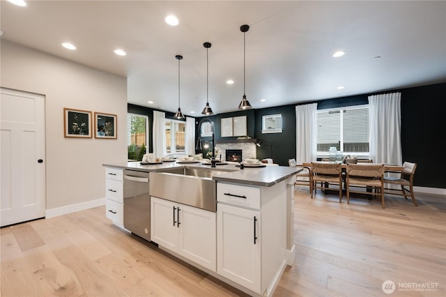 kitchen with stainless steel dishwasher, white cabinets, a sink, an island with sink, and light wood-type flooring