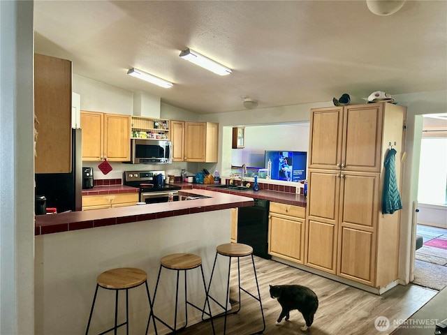 kitchen with light brown cabinets, a peninsula, a breakfast bar, light wood-style floors, and appliances with stainless steel finishes