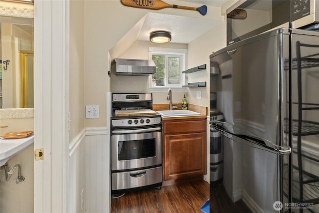 kitchen featuring stainless steel gas range, freestanding refrigerator, a sink, wall chimney exhaust hood, and brown cabinets