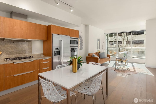 kitchen with stainless steel appliances, decorative backsplash, brown cabinetry, wood finished floors, and under cabinet range hood