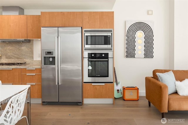 kitchen with under cabinet range hood, appliances with stainless steel finishes, light wood-type flooring, decorative backsplash, and brown cabinets