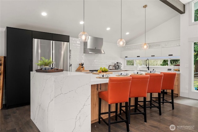 kitchen featuring dark wood-type flooring, wall chimney range hood, decorative backsplash, stainless steel refrigerator, and high vaulted ceiling