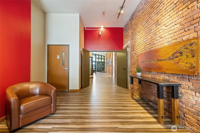 corridor with rail lighting, a towering ceiling, brick wall, wood finished floors, and baseboards