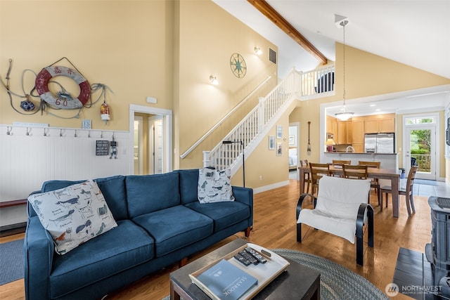 living area with high vaulted ceiling, a wainscoted wall, light wood-style floors, stairs, and beamed ceiling