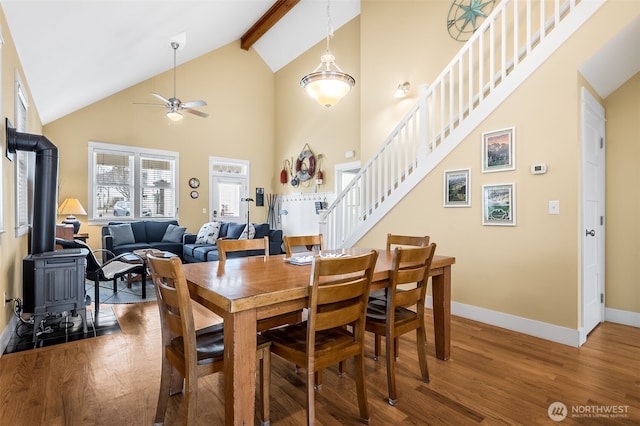 dining room featuring beam ceiling, a wood stove, wood finished floors, baseboards, and stairs