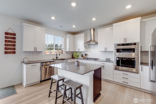 kitchen featuring light wood-style flooring, stainless steel appliances, a sink, a kitchen breakfast bar, and wall chimney exhaust hood