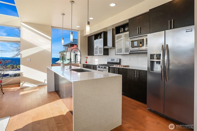 kitchen with a kitchen island with sink, a sink, wood finished floors, appliances with stainless steel finishes, and wall chimney range hood