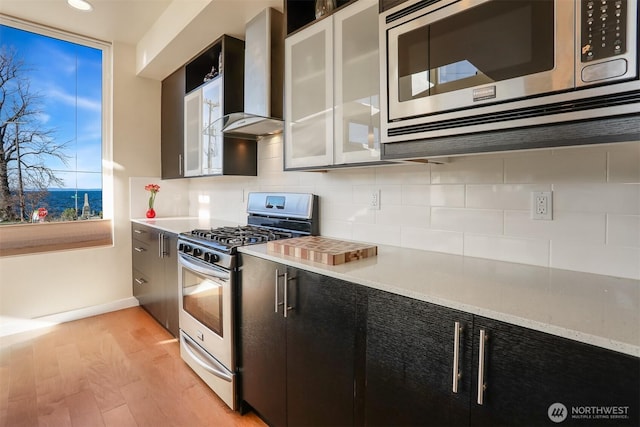 kitchen featuring stainless steel appliances, decorative backsplash, glass insert cabinets, wall chimney range hood, and light wood-type flooring