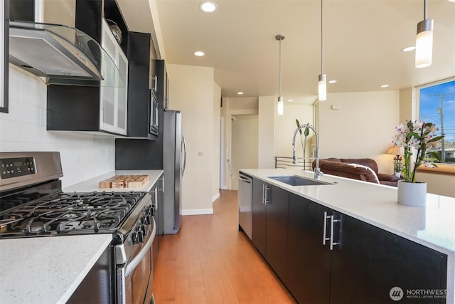 kitchen with light wood finished floors, wall chimney exhaust hood, appliances with stainless steel finishes, dark cabinetry, and a sink