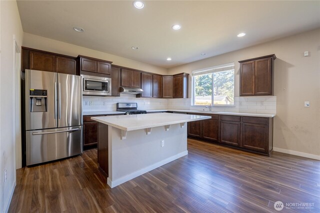 kitchen with dark wood-type flooring, under cabinet range hood, backsplash, appliances with stainless steel finishes, and dark brown cabinets