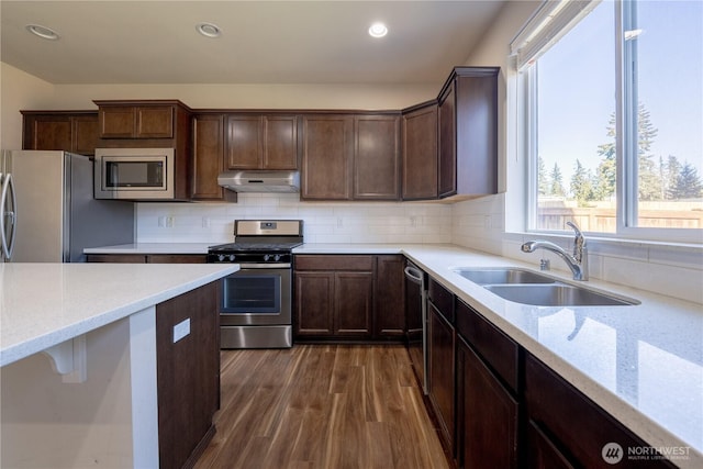 kitchen with a sink, under cabinet range hood, decorative backsplash, appliances with stainless steel finishes, and dark wood-style flooring