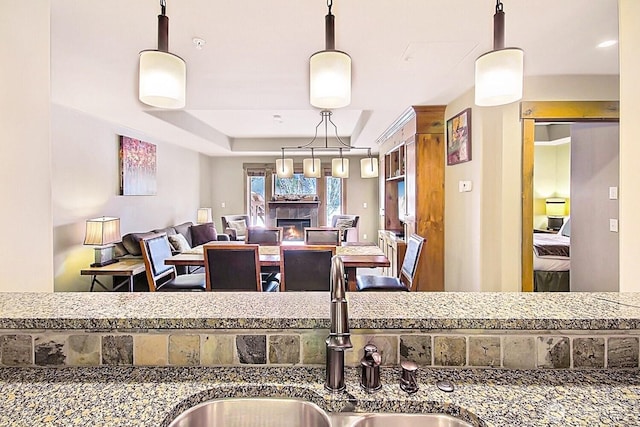 interior space with a glass covered fireplace, hanging light fixtures, a tray ceiling, stone counters, and a sink