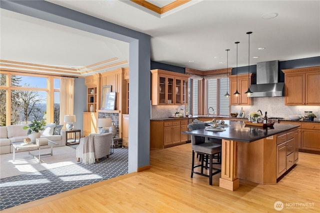 kitchen featuring a raised ceiling, dark countertops, wall chimney exhaust hood, open floor plan, and a kitchen bar