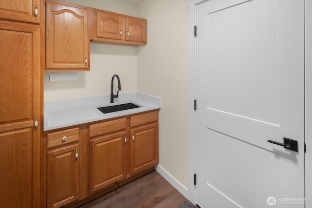 kitchen featuring light countertops, dark wood-type flooring, brown cabinetry, a sink, and baseboards