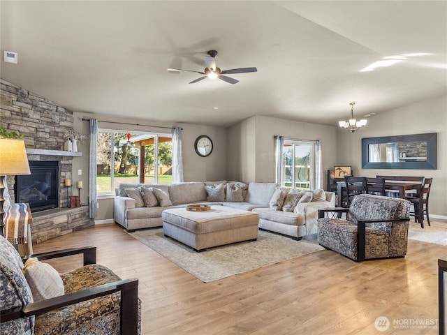 living room with a stone fireplace, light wood-style flooring, ceiling fan with notable chandelier, baseboards, and vaulted ceiling