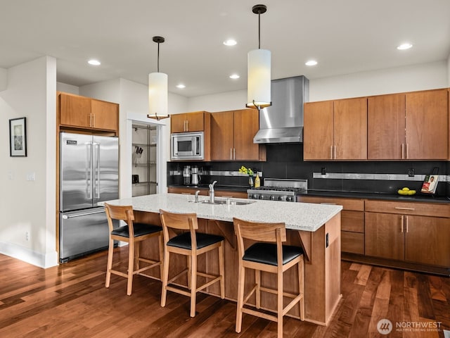 kitchen featuring wall chimney range hood, brown cabinetry, a sink, and built in appliances