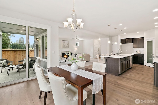 dining room featuring a warm lit fireplace, ceiling fan with notable chandelier, light wood finished floors, and recessed lighting