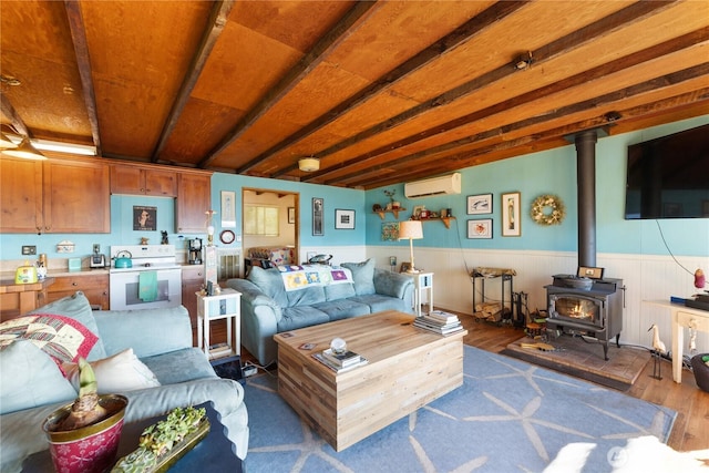 living room featuring wood finished floors, a wainscoted wall, a wood stove, an AC wall unit, and beamed ceiling