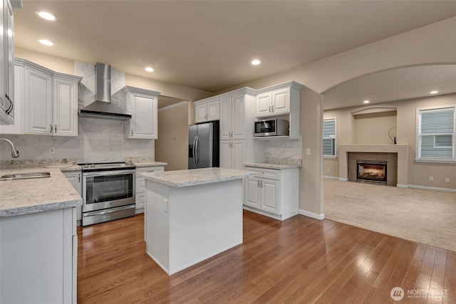 kitchen with stainless steel appliances, a sink, open floor plan, wall chimney range hood, and a glass covered fireplace