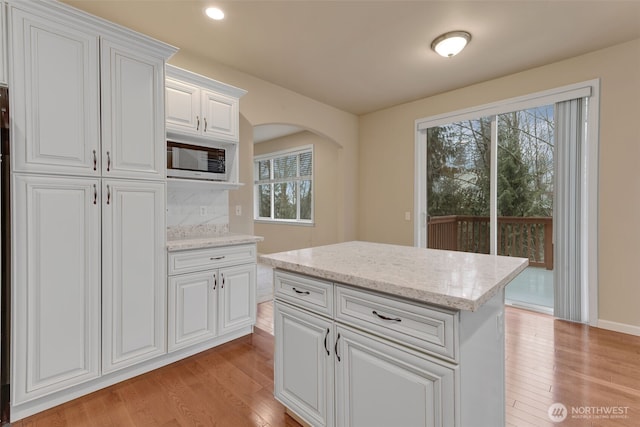 kitchen with arched walkways, white cabinets, light wood-type flooring, light stone countertops, and stainless steel microwave